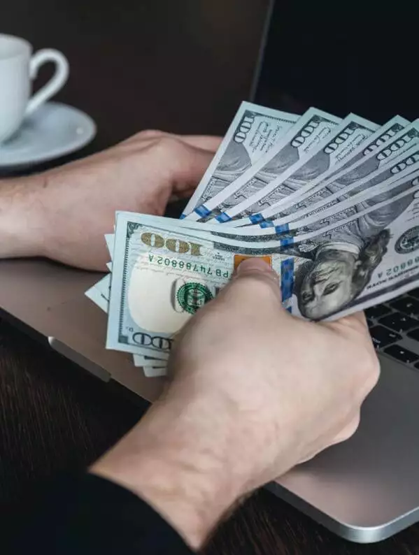 a person at a table counting u.s. dollars near an open laptop and a coffee cup, exploring ways to make money from their blog.