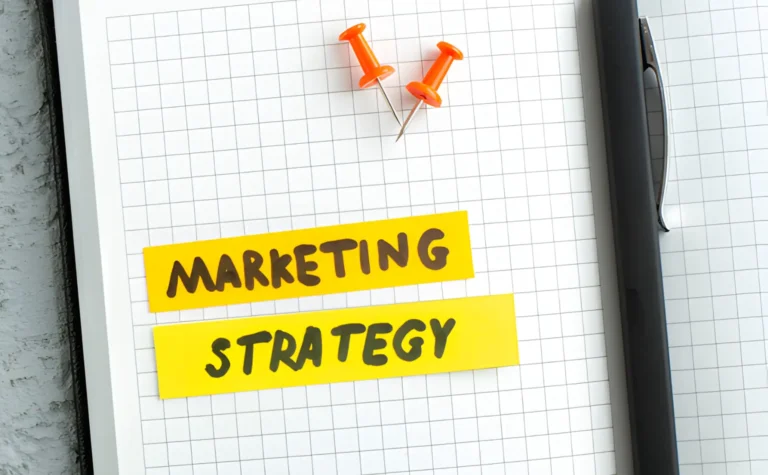 a notepad with a grid pattern features two yellow sticky notes labeled "marketing strategy" in black. two red pushpins and a black pen are placed nearby, ready to help you propel your business over the next 12 months.