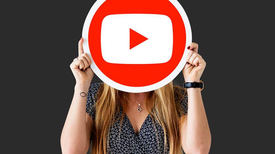a woman holds a large youtube play button sign in front of her face against a dark background, symbolizing her journey as a full-time youtuber.
