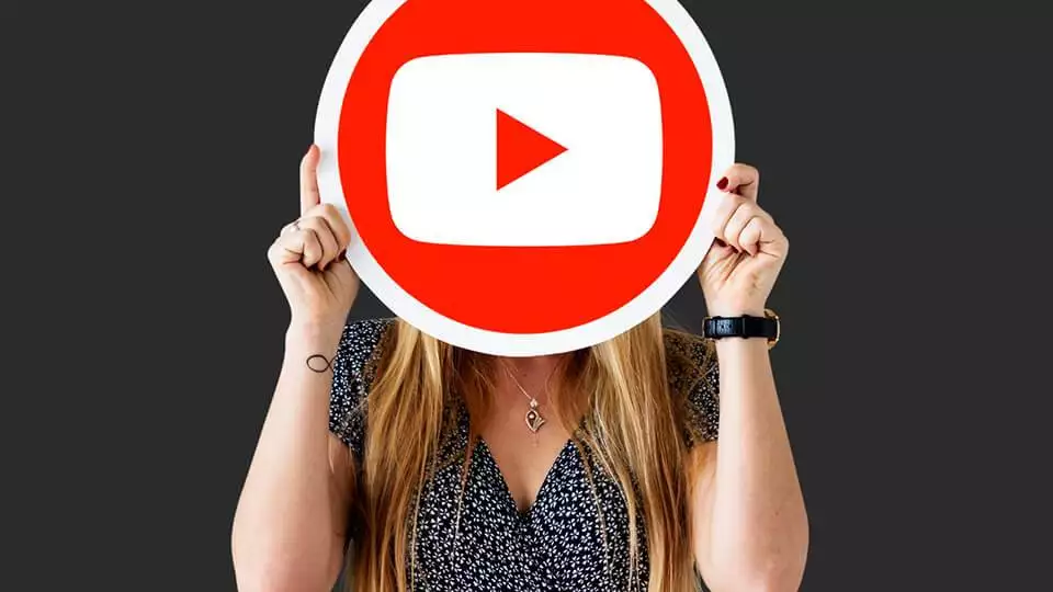 a woman holds a large youtube play button sign in front of her face against a dark background, symbolizing her journey as a full-time youtuber.