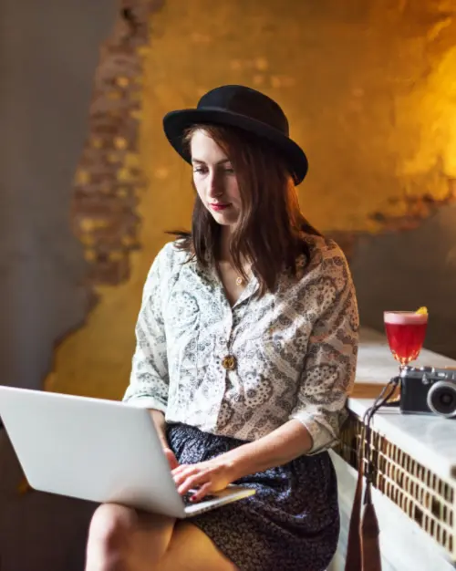 A woman wearing a hat uses a laptop while sitting at a counter with an exposed brick wall, a camera, and a cocktail nearby, possibly brainstorming business ideas for a niche market.