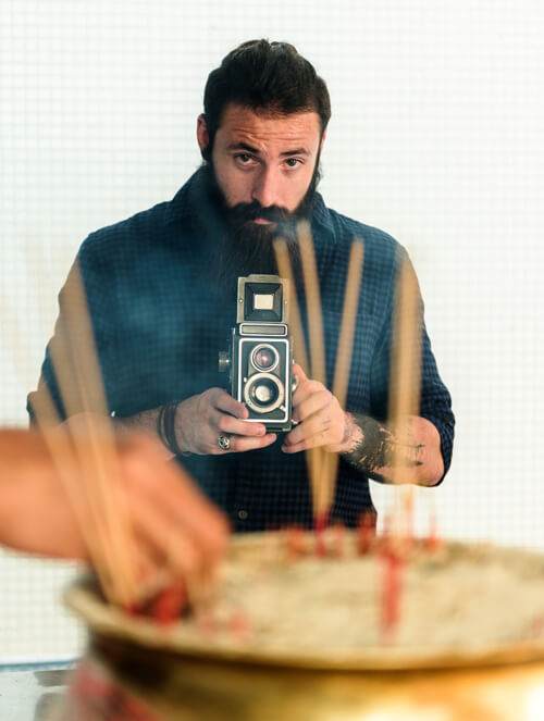 a man with a beard holding a vintage camera, focusing intently, with a blurred foreground of candles on a cake, capturing the essence of social media authenticity.