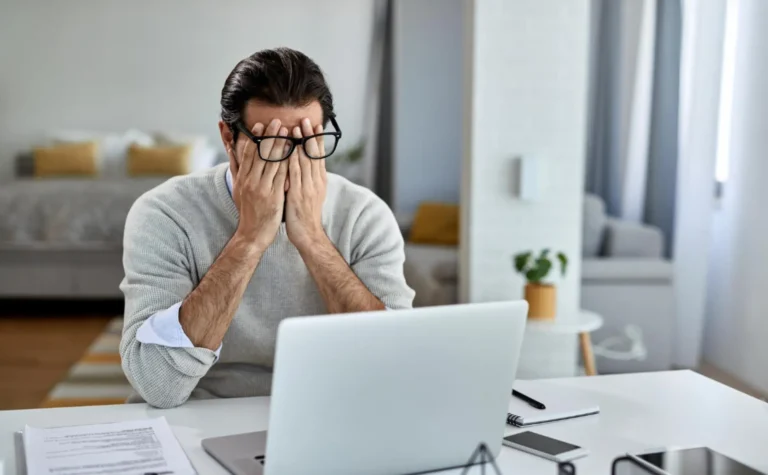 a person sits at a desk with their hands covering their face in front of a laptop, appearing stressed or tired due to unexpected challenges. a document is on the table, and a bed and plant are visible in the background.