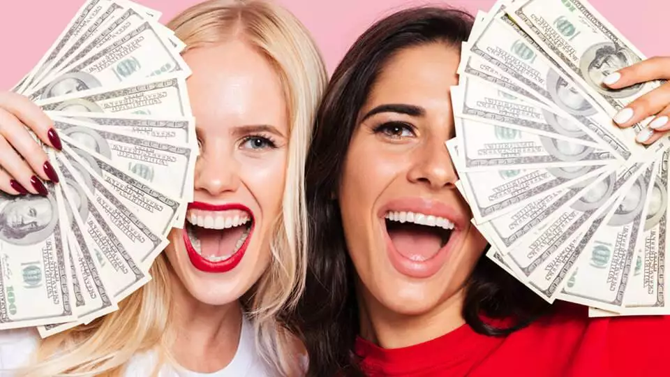 two joyful women holding multiple 100 dollar bills against a pink background, celebrating their success in doubling their online income.